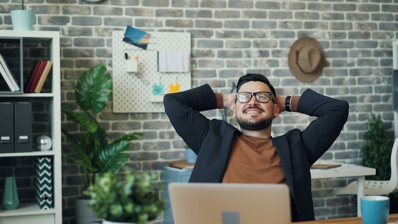 Man leaning back from his laptop with his hands behind his head, taking a break at his desk